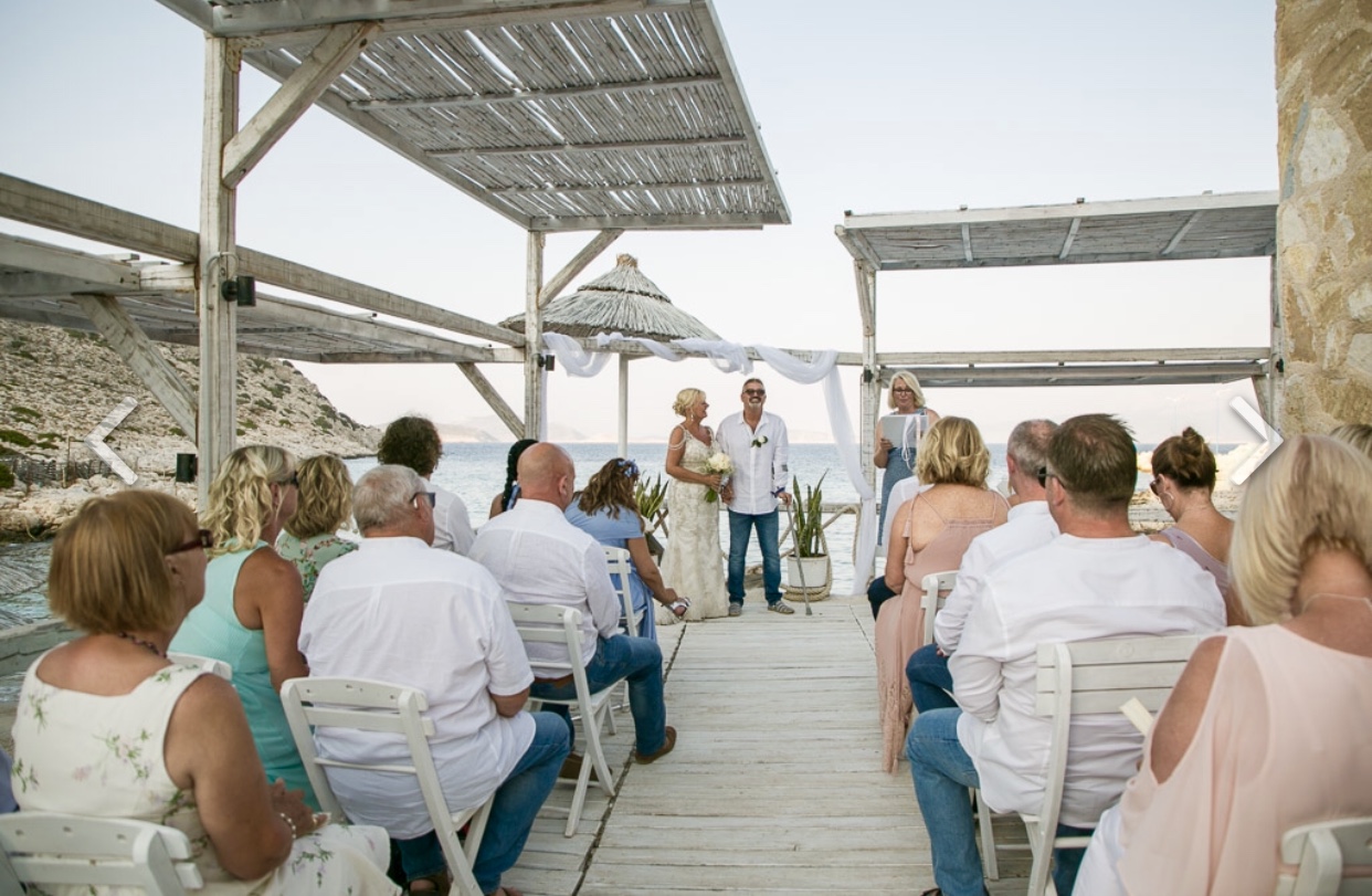 A Greek beach wedding ceremony in Halki, Greece. The photo shows the guests seated and the couple in the bckground with the Celebrant and a view of the sea.